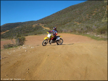 Man wearing black and red motocross gear with helmet riding Suzuki RM-250 in dirt parking lot.