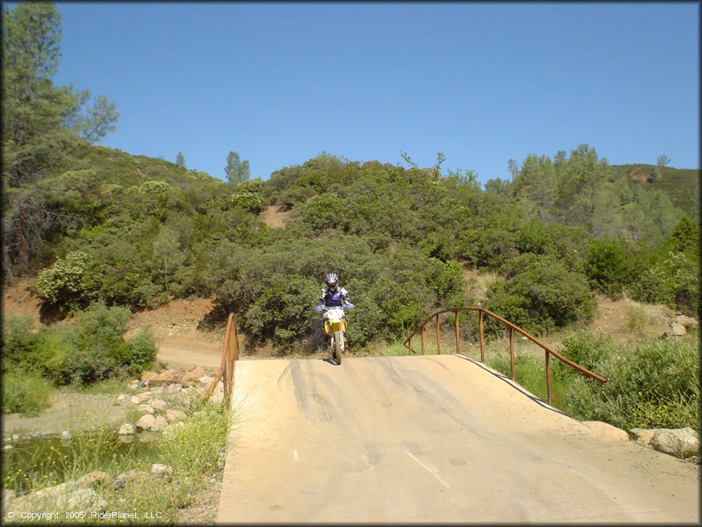 Young woman on Suzuki dirt bike riding over small bridge crossing.