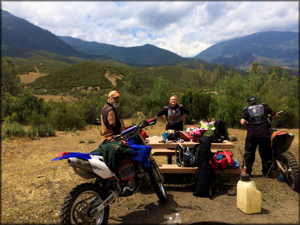 Three people standing next to picnic table with pile of motorcycle riding gear, a gasoline can and KTM and Yamaha dirt bike.